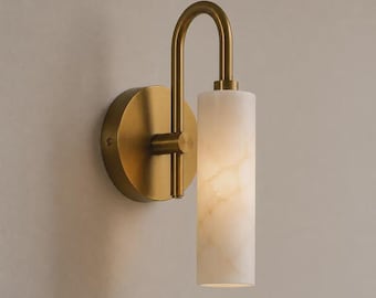 May include: A wall sconce featuring a circular brass mounting plate and a curved brass arm that supports a cylindrical white stone shade with natural veining. The light fixture has a warm metallic finish and a vertical, tubular design.