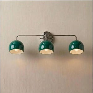 May include: A wall-mounted light fitting featuring a horizontal metallic bar with three evenly spaced, dome-shaped shades in a glossy dark green finish. Each shade has a white interior and emits a warm glow from the light bulb inside.