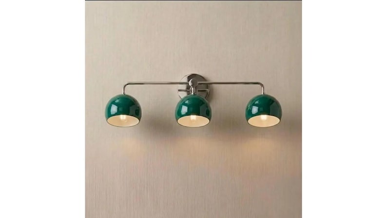 May include: A wall-mounted light fitting featuring a horizontal metallic bar with three evenly spaced, dome-shaped shades in a glossy dark green finish. Each shade has a white interior and emits a warm glow from the light bulb inside.