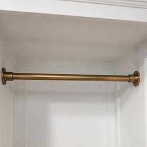 Puede incluir: Una barra de armario horizontal de latón con un acabado metálico liso. La barra cuenta con bridas de montaje circulares en ambos extremos, cada una asegurada con tres tornillos visibles, diseñada para su instalación entre dos paneles blancos.