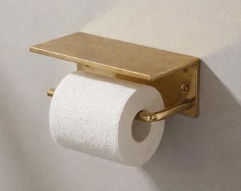 May include: A wall-mounted toilet roll holder made of brushed brass with a flat rectangular shelf on top. The holder features a horizontal bar that supports a single roll of white toilet paper with a textured, embossed pattern.