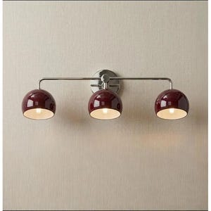 Pode incluir: Um candeeiro de parede de três luzes com uma barra horizontal em cromo polido e um suporte circular. Três cúpulas brilhantes em tom bordeaux profundo estão fixadas à barra, cada uma com um acabamento interior na cor branca.