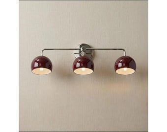 Puede incluir: Un aplique de pared de tres luces con una barra horizontal de cromo pulido y un soporte circular. Tres pantallas brillantes en forma de cúpula de color burdeos intenso están unidas a la barra, cada una con un acabado interior blanco.