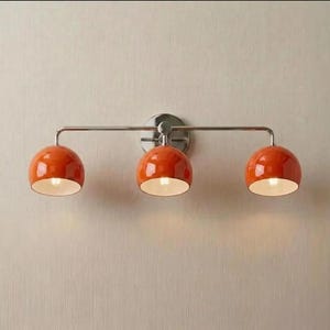 May include: A wall-mounted light fitting featuring a horizontal brushed nickel bar and a circular backplate. Three glossy orange dome-shaped shades are attached to the bar, each housing a single light bulb with a white interior finish.