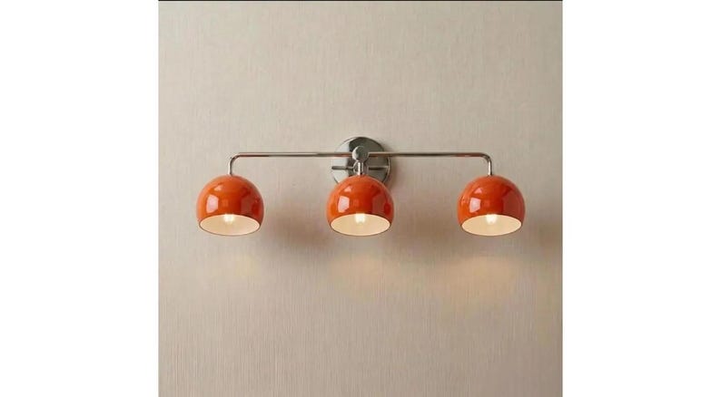 May include: A wall-mounted light fitting featuring a horizontal brushed nickel bar and a circular backplate. Three glossy orange dome-shaped shades are attached to the bar, each housing a single light bulb with a white interior finish.