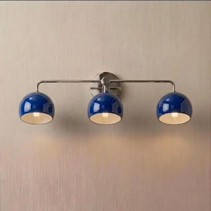May include: A wall-mounted light fitting featuring a horizontal metallic bar with three evenly spaced, glossy blue dome-shaped shades. Each shade points downwards, revealing a white interior and a glowing light bulb inside the fitting.