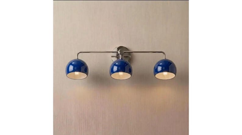 May include: A wall-mounted light fitting featuring a horizontal metallic bar with three evenly spaced, glossy blue dome-shaped shades. Each shade points downwards, revealing a white interior and a glowing light bulb inside the fitting.