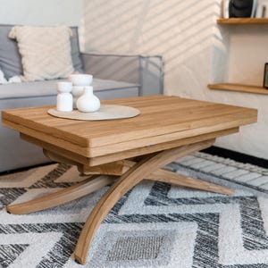 Op de afbeelding: Een rechthoekige lichtbruine houten salontafel met een dik blad en gebogen, kruisende poten. De tafel heeft een gladde afwerking met een natuurlijke houtnerf en een stevig onderstel. Drie kleine witte decoratieve vazen staan op een ronde mat.