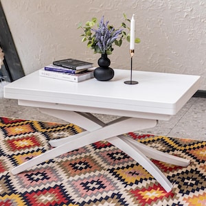 Op de afbeelding: Een witte rechthoekige salontafel met een kruisvormig onderstel, voorzien van een stapel van twee boeken, een zwarte vaas met paarse lavendelbloemen en een lange witte kaars in een zwarte houder op het gladde, vlakke tafeloppervlak.