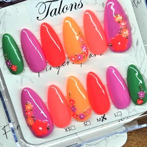 May include: A set of twelve almond-shaped press-on nails in vibrant neon colors including green, pink, and orange. Several nails feature small floral decals and gold-toned metallic studs. The nails are displayed on a white card labeled Talons.