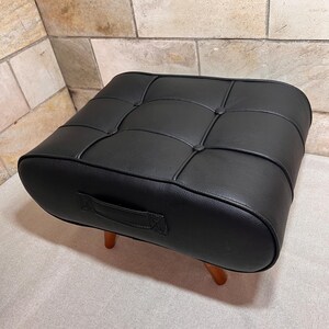 May include: A black rectangular ottoman featuring a tufted top with button accents and a side handle. The piece is supported by four short, tapered wooden legs, providing a stable base for this functional piece of furniture.