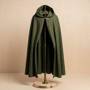 Puede incluir: Una capa con capucha de color verde oliva que llega hasta el suelo, con un acabado de tela texturizada. La prenda se sujeta al cuello con una correa de cuero marrón y un cierre de hebilla metálica, cayendo desde los hombros en una silueta amplia.