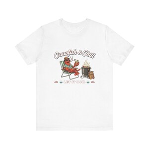 Puede incluir: Una camiseta blanca con un gráfico de un cangrejo de río rojo con sombrero y gafas de sol, sentado en una silla plegable con una bebida. Al lado hay una olla humeante. El texto dice Crawfish & Chill y LET IT BOIL.