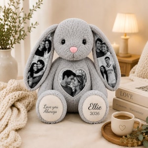 Könnte beinhalten: Ein graues Plüschkaninchen mit einer rosa Nase und langen Ohren. Die Ohren und die Brust zeigen schwarz-weiße Fotodrucke von Personen. Die Füße des Kaninchens sind mit dem Text Love you Always und Ellie 2026 bestickt.