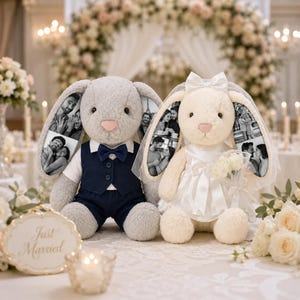 May include: Two plush rabbit dolls dressed for a wedding. The gray rabbit wears a navy blue vest and bow tie. The cream rabbit wears a white satin dress, a veil, and holds a small bouquet. Both rabbits feature black and white photo prints on their ears.
