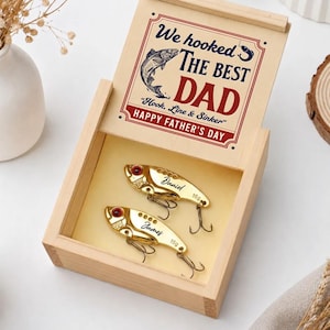 May include: A light-colored wooden box with a sliding lid featuring the text "We hooked THE BEST DAD Hook, Line & Sinker HAPPY FATHER'S DAY" and a fish graphic. Inside are two gold-colored metal fishing lures, each engraved with a name and 15g.