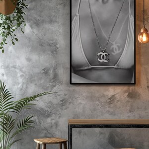 May include: A black and white framed wall art print featuring a close-up of a bikini top and a silver-toned necklace with a prominent interlocking double C logo pendant. The artwork is displayed in a thin black frame, capturing the metallic sheen of the jewelry and the fabric texture of the swimwear in a high-contrast monochromatic composition.