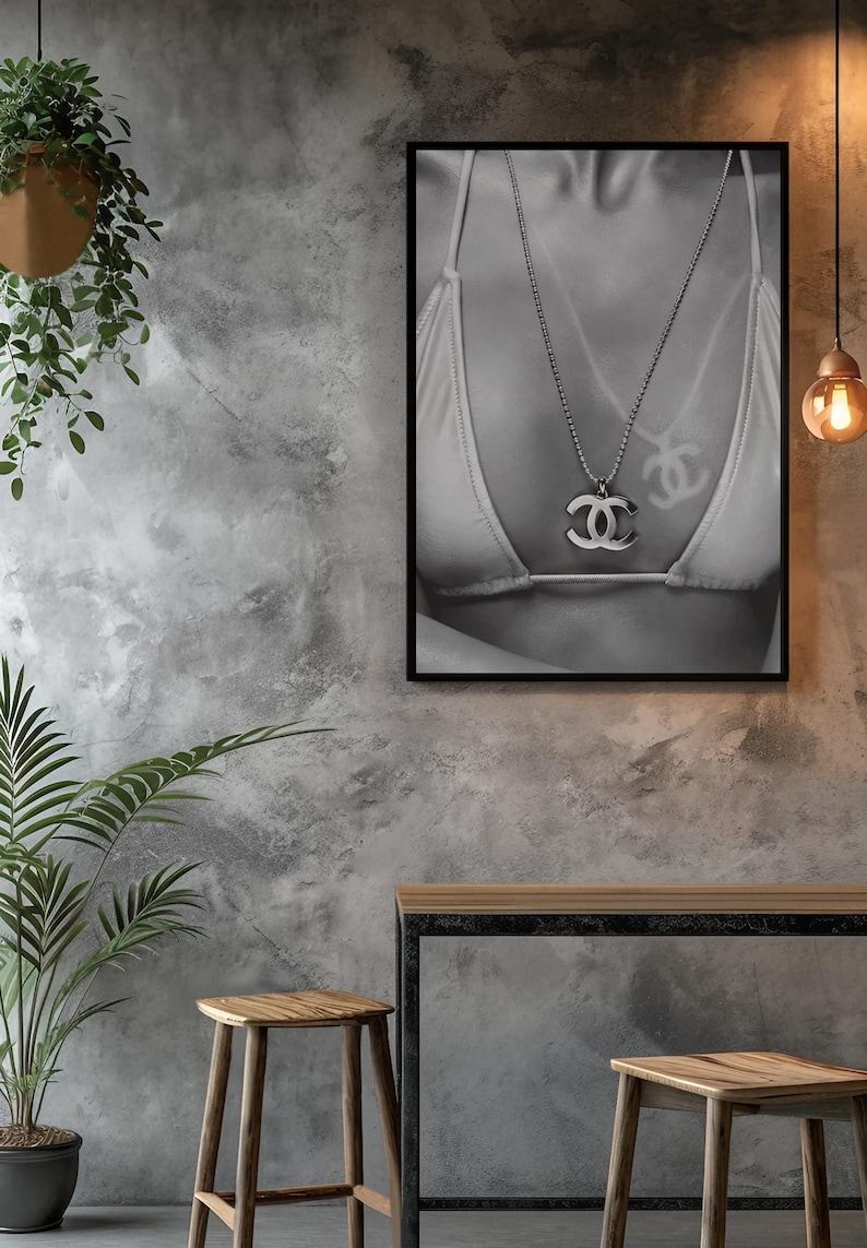 May include: A black and white framed wall art print featuring a close-up of a bikini top and a silver-toned necklace with a prominent interlocking double C logo pendant. The artwork is displayed in a thin black frame, capturing the metallic sheen of the jewelry and the fabric texture of the swimwear in a high-contrast monochromatic composition.