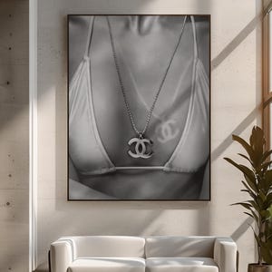 May include: A black and white wall art print featuring a close-up of a person wearing a bikini top and a silver-toned chain necklace with a prominent interlocking CC logo pendant. The image displays a soft shadow of the logo cast on the fabric.