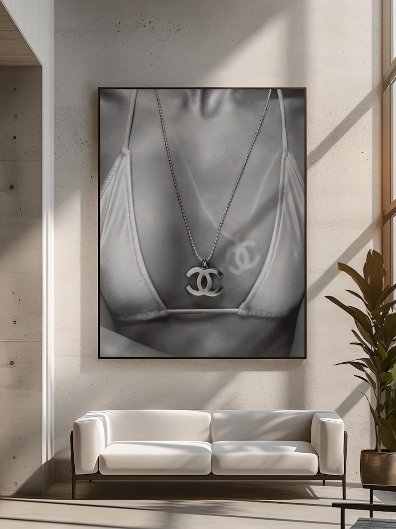 May include: A black and white wall art print featuring a close-up of a person wearing a bikini top and a silver-toned chain necklace with a prominent interlocking CC logo pendant. The image displays a soft shadow of the logo cast on the fabric.