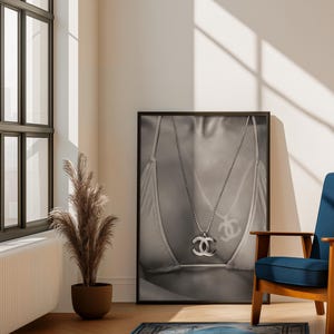May include: A black and white framed wall art print featuring a close-up of a person wearing a bikini top and a silver-toned chain necklace with a prominent interlocking CC logo pendant. The image captures the metallic texture of the jewelry.