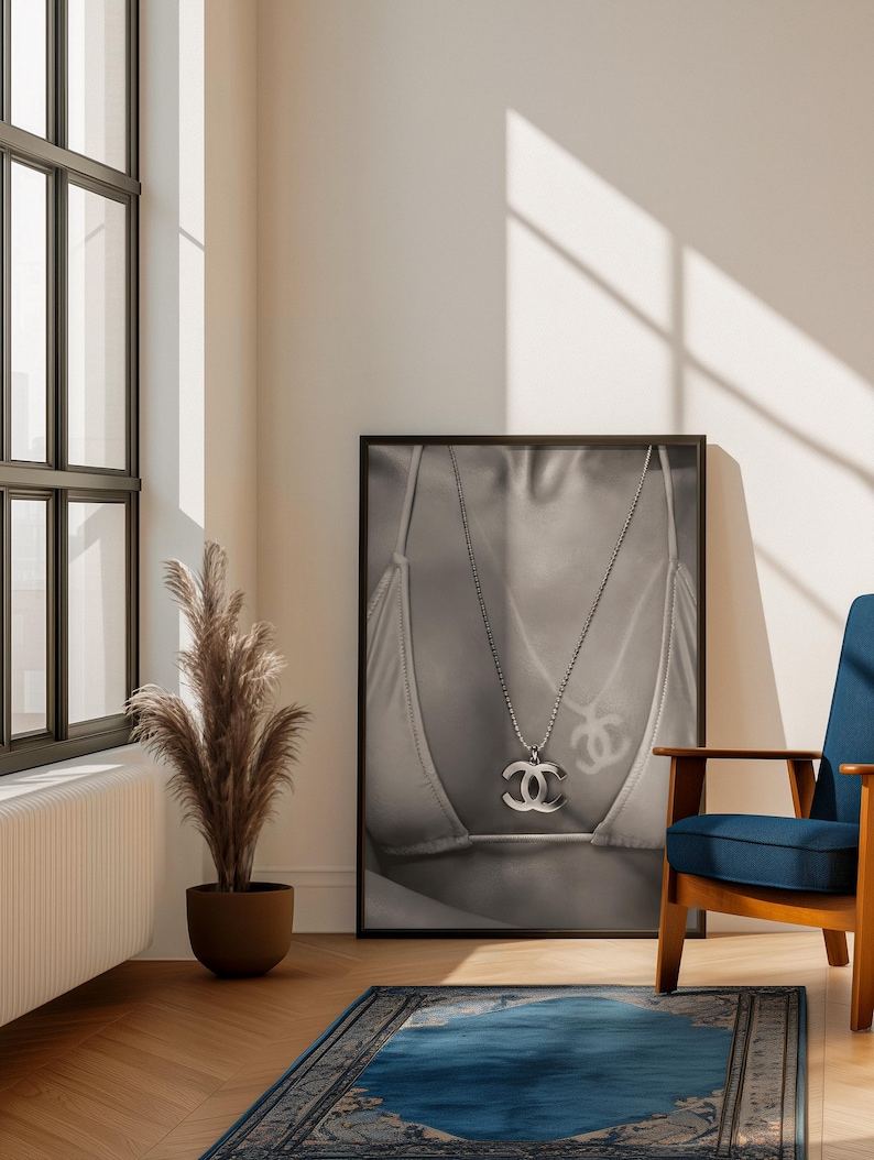 May include: A black and white framed wall art print featuring a close-up of a person wearing a bikini top and a silver-toned chain necklace with a prominent interlocking CC logo pendant. The image captures the metallic texture of the jewelry.