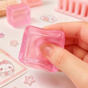 May include: A translucent, squishy, pink cube made of a soft, flexible material. The cube has a smooth, glossy surface with a subtle pearlescent sheen throughout its structure, allowing light to pass through its semi-transparent, light pink body.