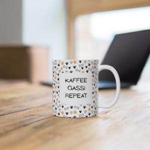 Könnte beinhalten: Ein weißer Keramikbecher mit einem wiederkehrenden Muster aus schwarzen Pfotenabdrücken, kleinen Herzen, Knochen und orangefarbenen Tennisbällen. In der Mitte steht der schwarze Text "KAFFEE GASSI REPEAT" mit einem Henkel auf der rechten Seite.