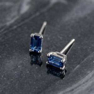Puede incluir: Un par de pendientes de botón con gemas azules rectangulares de corte esmeralda engastadas en metal plateado pulido. Cada pendiente tiene un poste fino y una superficie facetada de color azul profundo y claro que refleja la luz.