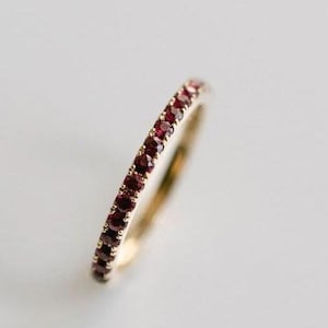 Puede incluir: Un anillo de banda fina de oro con una hilera continua de pequeñas piedras preciosas redondas de color rojo intenso engastadas a lo largo de la superficie exterior. El metal tiene un acabado pulido y las piedras forman un patrón circular.