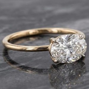 Puede incluir: Un anillo de oro con una piedra preciosa transparente de corte ovalado engastada en cuatro garras. La banda es lisa y pulida, mostrando un diseño sencillo que resalta la piedra facetada. El anillo se muestra desde un ángulo lateral.