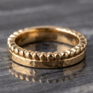 Puede incluir: Un anillo de oro con un diseño de doble banda. La parte superior consiste en una fila continua de pequeñas cuentas esféricas en relieve, mientras que la parte inferior es una banda plana y lisa con una textura martillada en la superficie.