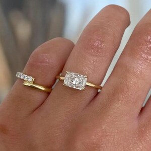 Puede incluir: Dos anillos de oro en los dedos. Uno presenta una piedra clara de corte rectangular montada horizontalmente en una banda fina de oro. El segundo es un anillo de estilo abierto con una hilera de pequeñas piedras claras de corte redondo.