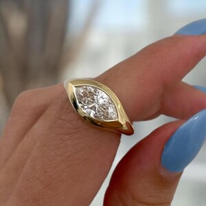 Puede incluir: Un anillo de oro con una piedra preciosa transparente de corte marquesa engastada en un bisel liso y pulido. La banda de oro macizo presenta un diseño cónico que enmarca la forma alargada y puntiaguda de la piedra central, creando un aspecto elegante.