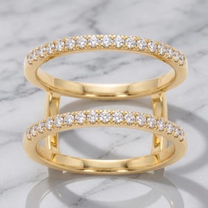 Puede incluir: Un anillo de doble banda en oro con dos bandas paralelas conectadas por barras verticales de oro. Cada banda está engastada con una fila de pequeñas piedras claras de corte redondo que cubren la mitad superior, creando un aspecto metálico.