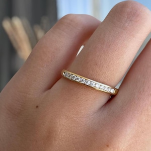 Puede incluir: Un anillo de banda dorada con un engaste de canal que presenta una hilera de pequeñas piedras redondas y transparentes incrustadas en el centro. El metal tiene un acabado pulido y reflectante, con las piedras alineadas en la parte superior.