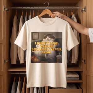 Könnte beinhalten: Ein cremefarbenes Kurzarm-T-Shirt mit einer Grafik einer runden, braun-weißen Katze, die auf einem Sofa sitzt. Der gelbe Text lautet: I DIDN'T LOSE WEIGHT BECAUSE I NEVER LOSE. Die Katze befindet sich neben einer Tüte Chips und einem Getränk.