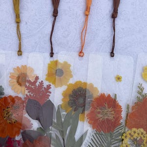 May include: A set of four clear rectangular bookmarks featuring pressed flowers and foliage in shades of yellow, orange, and brown. Each bookmark is topped with a matching colored thread tassel in mustard, dark brown, or bright orange.