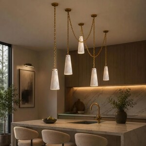 May include: A multi-light pendant fixture featuring five cone-shaped white marble shades suspended by gold-toned chains. The chains are draped in a swag pattern from the ceiling, connecting the individual lights above a kitchen island.
