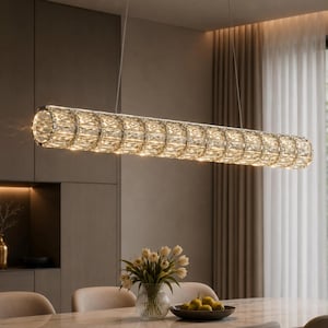 Pode incluir: Uma luminária pendente cilíndrica e longa suspensa por fios finos. O exterior é composto por numerosos segmentos transparentes e facetados, dispostos num padrão repetitivo que refrata a luz, criando um aspeto brilhante e cintilante.
