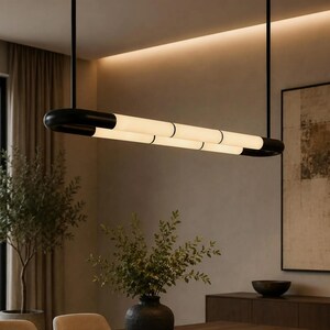 May include: A linear pendant light fixture featuring a matte black metal frame with two parallel, horizontal, cylindrical frosted glass tubes. The fixture is suspended from the ceiling by two thin black rods, emitting a soft, warm glow.