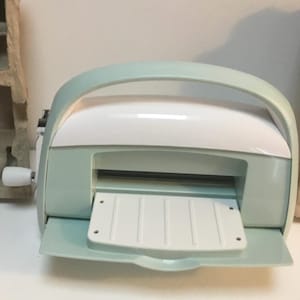 May include: A light mint green and white manual die-cutting and embossing machine featuring a curved top handle, a central slot for material insertion, a fold-down front platform with vertical ridges, and a white hand crank on the left side.