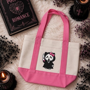 May include: A cream-colored canvas tote bag with bright pink handles and a pink base. The front features a graphic of a black-robed character wearing a white mask, holding a knife, and sporting a pink bow on its head, centered on the bag.