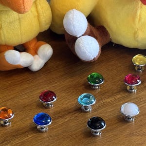 May include: A collection of colorful, decorative cabinet knobs with clear, jewel-like tops in various colors, including orange, red, blue, green, yellow, and white. Each knob has a silver-colored base. The knobs are arranged on a wooden surface.