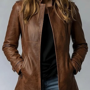 Puede incluir: Una chaqueta de cuero marrón con cuello alzado, mangas largas con detalles en los puños y dos bolsillos delanteros. La chaqueta se lleva sobre una camiseta negra y vaqueros azules, mostrando una textura suave y costuras visibles.