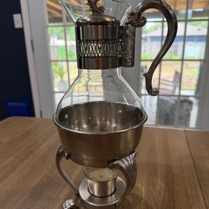 May include: A clear glass carafe with a silver-toned metal base, handle, and decorative collar. The carafe sits on a three-legged metal stand that features a central holder containing a small white tea light candle for warming the contents.