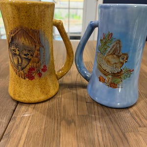 May include: Two ceramic mugs with handles. The left mug is speckled yellow and features an illustration of two raccoons inside a tree hollow. The right mug is light blue and features an illustration of a squirrel perched on a tree stump.