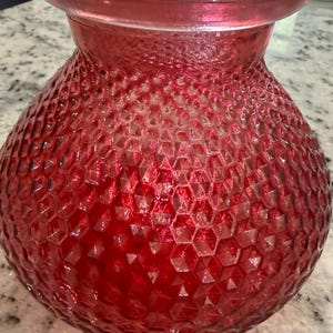 May include: A translucent red glass vase with a rounded, bulbous body and a wide, flared rim. The exterior surface features a repeating, textured honeycomb or diamond-shaped pattern that covers the entire rounded section of the vessel.