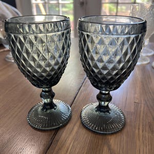 May include: Two matching stemmed goblets crafted from translucent dark gray glass. Each vessel features a prominent diamond-shaped geometric pattern across the bowl, a smooth neck, and a circular base with a radiating ribbed texture.