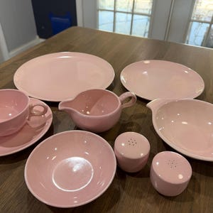 Op de afbeelding: Een set lichtroze keramieken servies bestaande uit twee ronde borden, een theekopje met bijpassende schotel, een kleine melkkan, een kom, een grotere ovale serveerschaal en twee kleine cilindervormige zout- en pepervaatjes.